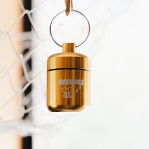 Könnte beinhalten: Ein zylindrischer, goldfarbener Metallbehälter mit Schraubdeckel und einem silberfarbenen Metallschlüsselring an der Oberseite. Die Seite des Behälters zeigt den eingravierten Text BEEHUG2 über einer kleinen Grafik einer schlafenden Biene.