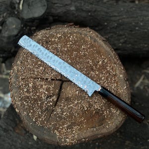 Könnte beinhalten: Ein langes Wellenschliff-Brotmesser mit einer gehämmerten Metallklinge, das eine markante wellenförmige kupferfarbene Linie entlang der Schneide aufweist, sowie einen dunklen, polierten Holzgriff in zylindrischer Form mit glatter Oberfläche.