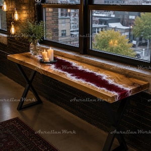 May include: A long rectangular console table featuring a natural wood slab top with a central deep red resin river inlay bordered by white accents. The table is supported by black metal X-shaped legs and is positioned in front of a large window.