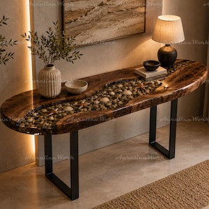 Könnte beinhalten: Ein ovaler Konsolentisch mit einer dunklen Holzplatte in Naturkante und einer zentralen Einlage aus glatten Flusskieseln, Muscheln und Seesternen, getragen von zwei schwarzen U-förmigen Metallbeinen, dekoriert mit einer Lampe und Schalen.