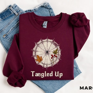 May include: A maroon crewneck sweatshirt featuring a graphic of a spider web with a spider, autumn leaves, and red berries. The text "Tangled Up" is printed in white below the graphic. The sweatshirt is layered over blue denim jeans.