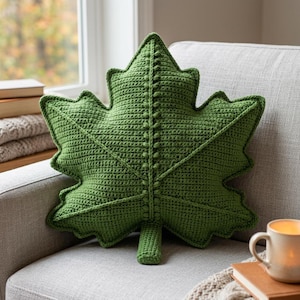May include: A green leaf-shaped throw cushion crafted with a crochet texture. The cushion features a central stem detail with small raised bobble stitches running vertically and horizontally across the surface, mimicking the veins of a leaf.