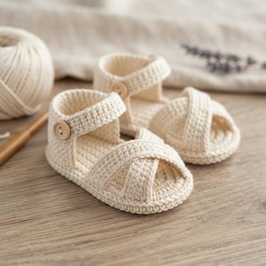 May include: A pair of cream-coloured crocheted sandals featuring a crisscross strap design over the toe area, an ankle strap with a single wooden button closure, and a textured knit construction made from thick yarn, presented as a matching set.