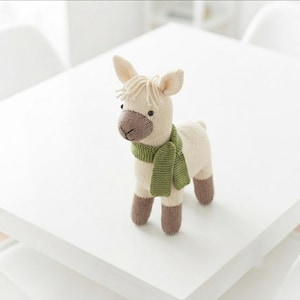 May include: A cream-colored knitted llama toy featuring a brown muzzle, dark button eyes, and a tuft of yarn hair on its head. The llama wears a textured green knitted scarf around its neck and has brown knitted feet on all four legs.
