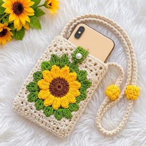 May include: A cream-colored crochet phone pouch featuring a large, three-dimensional sunflower motif with yellow petals, a brown center, and green leaves. The pouch has a long matching strap and a single white pearl button closure at the top.
