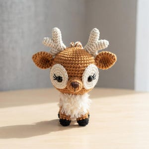 May include: A small, crocheted deer figurine featuring a brown head and body, cream-colored antlers, and white patches around the black eyes. The deer has a fluffy white chest, a dark brown nose, and small black hooves at the base of its body.