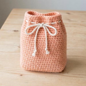 May include: A peach-coloured crochet drawstring pouch featuring a textured stitch pattern and a thick white cord threaded through the top opening, tied into a bow at the front with two dangling ends that have small knotted tips at the bottom.