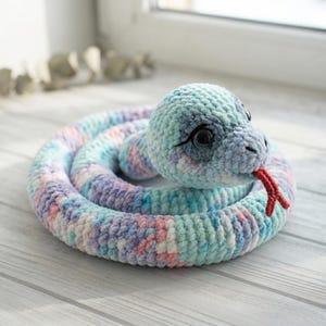 Puede incluir: Un juguete de serpiente de peluche enroscado, hecho de hilo suave y variado en tonos pastel de azul, rosa y blanco. La serpiente presenta ojos grandes, oscuros y brillantes con detalles cosidos en negro y una pequeña lengua roja bifurcada.