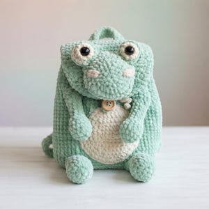 Puede incluir: Una mochila de ganchillo color verde menta con forma de rana, vientre color crema, dos grandes ojos de plástico negro y un pequeño cierre de botón de madera en el centro. La estructura de hilo texturizado incluye un asa y dos brazos.