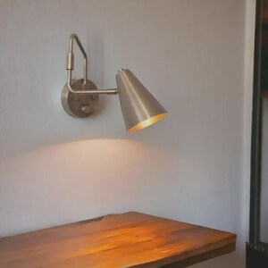 May include: A wall-mounted lamp with a brushed metallic finish, featuring a circular base plate and an adjustable arm that supports a conical shade. The lamp is angled downward, casting a warm, focused light onto the surface directly below it.