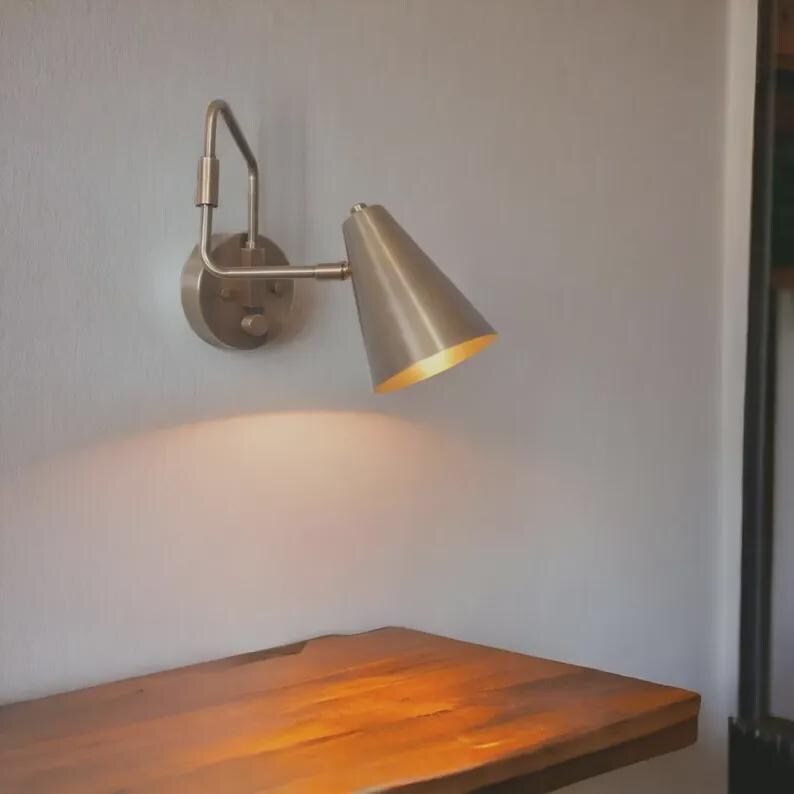 May include: A wall-mounted lamp with a brushed metallic finish, featuring a circular base plate and an adjustable arm that supports a conical shade. The lamp is angled downward, casting a warm, focused light onto the surface directly below it.
