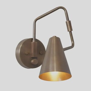 May include: A wall-mounted light fixture with a circular base and a conical shade. The lamp is constructed from a metallic material with a brushed bronze finish. A bent arm connects the base to the shade, which features a small knob on the side.