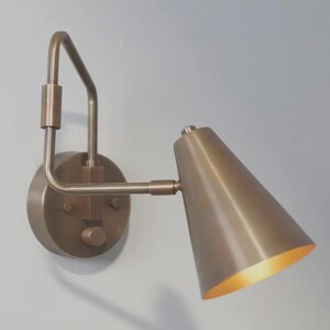 May include: A wall-mounted lamp with a metallic bronze finish, featuring a circular base plate, an angular arm, and a conical shade. The interior of the shade is a lighter, reflective gold tone, and a small circular knob is visible on the base.
