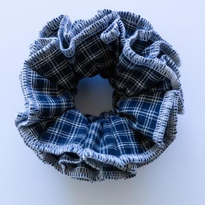 May include: A circular fabric hair scrunchie featuring a dark navy blue and white plaid pattern. The edges of the ruffled fabric are finished with a visible white serged stitch, creating a textured, gathered appearance around the elastic centre.