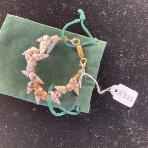 May include: A bracelet featuring small, spiraled, tan and white seashells interspersed with gold-toned beads and a gold-toned cylindrical clasp. A white rectangular tag attached to the bracelet displays the handwritten text #B25 in black ink.