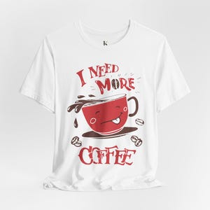 Può includere: Una t-shirt bianca a maniche corte con una grafica rossa di una tazza di caffè sorridente e il testo I NEED MORE COFFEE. Il design include chicchi di caffè rosso scuro e liquido che fuoriesce dalla tazza, centrato sulla parte anteriore del capo.