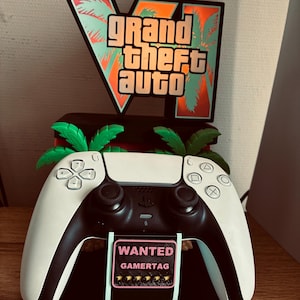 May include: A black and white video game controller resting on a custom display stand. The stand features a Grand Theft Auto V logo with palm trees and a rectangular plaque that reads WANTED GAMERTAG with five yellow stars underneath.