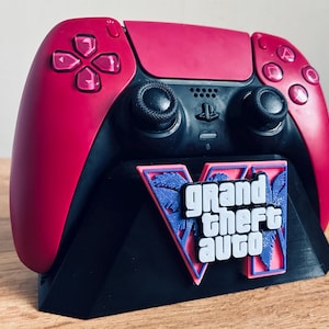 Può includere: Un controller per videogiochi magenta e nero appoggiato su un supporto espositivo nero. Il supporto presenta un logo Grand Theft Auto VI in bianco, rosa e blu, con una grafica di palma integrata nel design dietro la scritta.