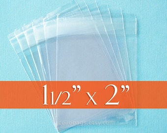 May include: Clear cellophane bags, 1.5" x 2", are stacked on a light blue surface. The bags are transparent and have a sealed top. The text "1 1/2" x 2"" is displayed on an orange banner.