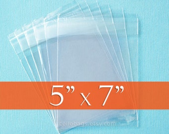 Könnte beinhalten: Klare Zellophanbeutel, 12,7 cm x 17,8 cm groß, gestapelt auf hellblauem Hintergrund. Die Beutel sind transparent und haben eine versiegelte Oberseite. Der Text "5" x 7"" ist weiß auf einem orangefarbenen Band dargestellt.