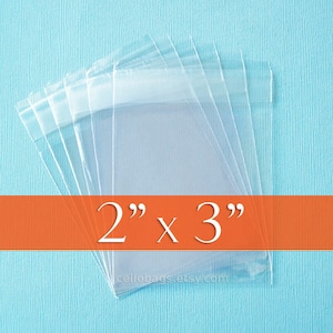 May include: Clear cellophane bags, 2" x 3", are stacked on a light blue surface. The bags are transparent and have a sealed top. The text "2" x 3"" is displayed in white on an orange banner.