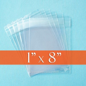 May include: Clear cellophane bags, each measuring 1 inch by 8 inches, are stacked on a light blue surface. The bags are transparent, allowing visibility of the contents. The text "1" x 8"" is displayed in white on an orange banner.