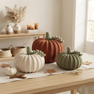 May include: Three crocheted pumpkins in cream, rust orange, and sage green, each featuring a ribbed texture and a green stem with a curly vine. A small white pumpkin and two cinnamon sticks are placed on a light-coloured table runner.