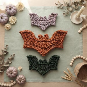 May include: Three crocheted bat appliques arranged vertically in a row. The top bat is light purple, the middle bat is burnt orange, and the bottom bat is dark green. Each bat features textured yarn stitches forming wings and ears.