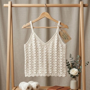 May include: A cream-colored sleeveless crochet top with a V-neckline and a scalloped hem, hanging on a wooden hanger. A brown paper tag attached to the hanger features the printed text V-STITCH TOP PATTERN in black ink on the front surface.
