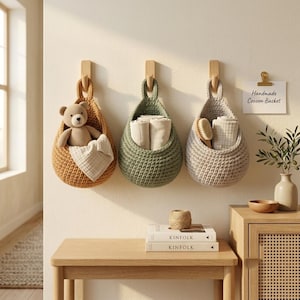 May include: Three hanging crochet baskets in mustard yellow, sage green, and beige, each suspended from a wooden hook. The baskets contain a teddy bear, folded white cloths, and a wooden brush. A sign reads Handmade Cocoon Basket.