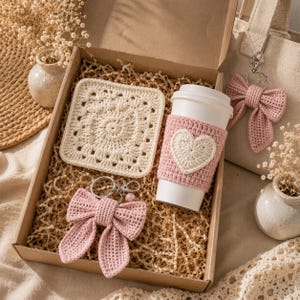 Könnte beinhalten: Eine Geschenkbox mit einem cremefarbenen, quadratischen Häkeluntersetzer, einem weißen Kaffeebecher mit einer rosafarbenen Häkelmanschette mit cremefarbenem Herz sowie zwei rosafarbenen Häkelschleifen-Schlüsselanhängern mit silberfarbenen Metallringen.