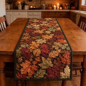 Puede incluir: Un camino de mesa rectangular con un patrón denso y repetitivo de hojas de otoño en tonos naranja, rojo, amarillo, marrón y verde apagado. El camino de tela tiene un borde verde oscuro y está centrado sobre una mesa de comedor de madera.