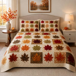 Pode incluir: Uma colcha e duas fronhas a condizer com um padrão de grelha de quadrados individuais, cada um exibindo uma folha de ácer em tons de vermelho, laranja, amarelo, castanho e verde sobre um fundo creme com costuras acolchoadas.