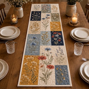 May include: A rectangular quilted table runner featuring a patchwork design of various wildflower illustrations in shades of blue, yellow, cream, and red. The fabric displays botanical prints of cornflowers, daisies, and lavender on a textured surface.