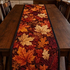 Puede incluir: Un camino de mesa rectangular con un diseño de tela acolchada que presenta un patrón denso de hojas de otoño caídas en tonos naranja, rojo, marrón y dorado. El camino tiene un borde de tela negra sólida y está centrado en una mesa de madera.