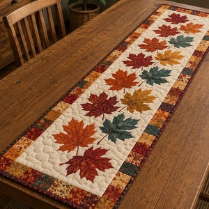 May include: A rectangular quilted table runner featuring a central cream-colored panel decorated with a repeating pattern of autumn maple leaves in shades of red, orange, yellow, and dark green, bordered by a patchwork frame of small squares.