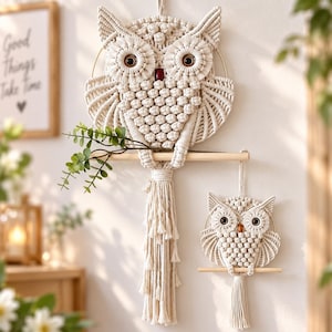 May include: Two off-white macrame owl wall hangings crafted from knotted cord. The larger owl features a long fringe and holds a green leafy branch on a wooden dowel. Both owls have dark brown circular eyes and a small red or brown bead nose.