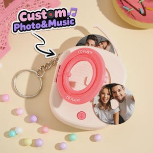 Puede incluir: Un llavero de reproductor de CD rosa claro con una inserción de foto circular de una pareja sonriente. El dispositivo tiene un centro circular rosa brillante con el texto CD Player impreso. Una cadena y un anillo de metal plateado están unidos.