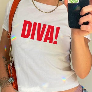 May include: A white short-sleeved t-shirt featuring the word DIVA! printed in bold red capital letters across the chest. The shirt is paired with a gold-toned chain necklace and an orange shoulder bag with a silver-toned metal clasp and chain.