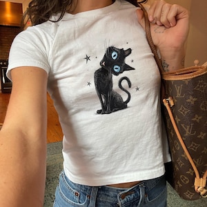May include: A white short-sleeved t-shirt featuring a graphic illustration of a black cat with large, bright blue eyes and a curved tail. The cat is surrounded by small black star shapes. The shirt has a crew neckline and a fitted silhouette.