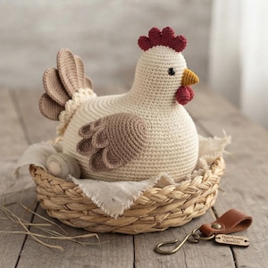 Può includere: Una gallina all'uncinetto color crema con ali e piume della coda marroni, cresta e bargigli rossi e becco giallo. La gallina è seduta in un cesto di paglia intrecciata foderato con tessuto bianco, accompagnata da un uovo di legno e un'etichetta.