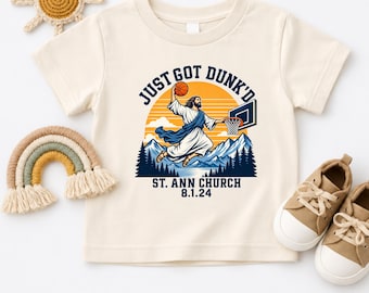 May include: A cream-colored short-sleeve t-shirt featuring a graphic of a figure in robes dunking a basketball. The text reads "JUST GOT DUNK'D" above the graphic and "ST. ANN CHURCH 8.1.24" below, set against a background of mountains and a sun.
