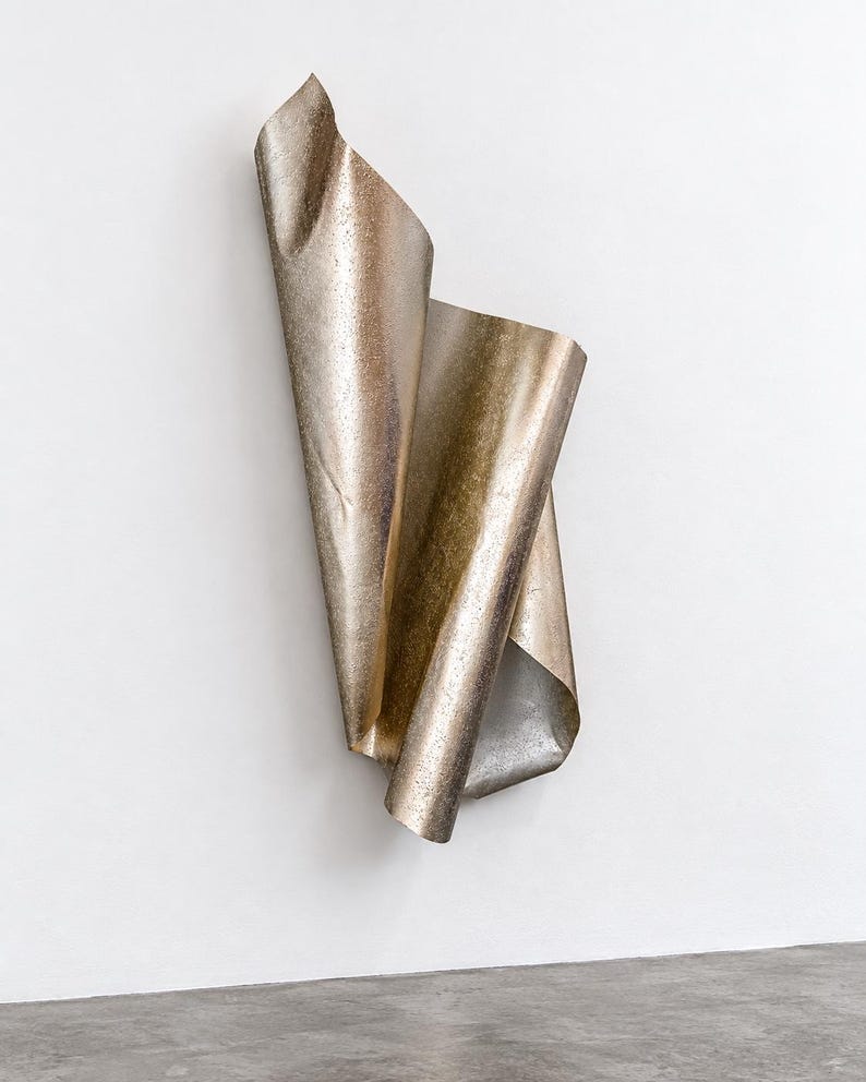 May include: A vertical wall sculpture made of a single sheet of metallic material with a textured, bronze-toned finish. The sheet is folded and curved into an elongated, flowing form that creates deep shadows and highlights along its surface.