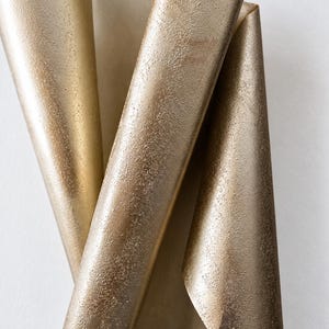 May include: A sheet of metallic gold paper with a textured, granular surface, folded into a series of vertical, overlapping curves that create a dynamic, three-dimensional shape with varying light reflections across the uneven, shimmering finish.