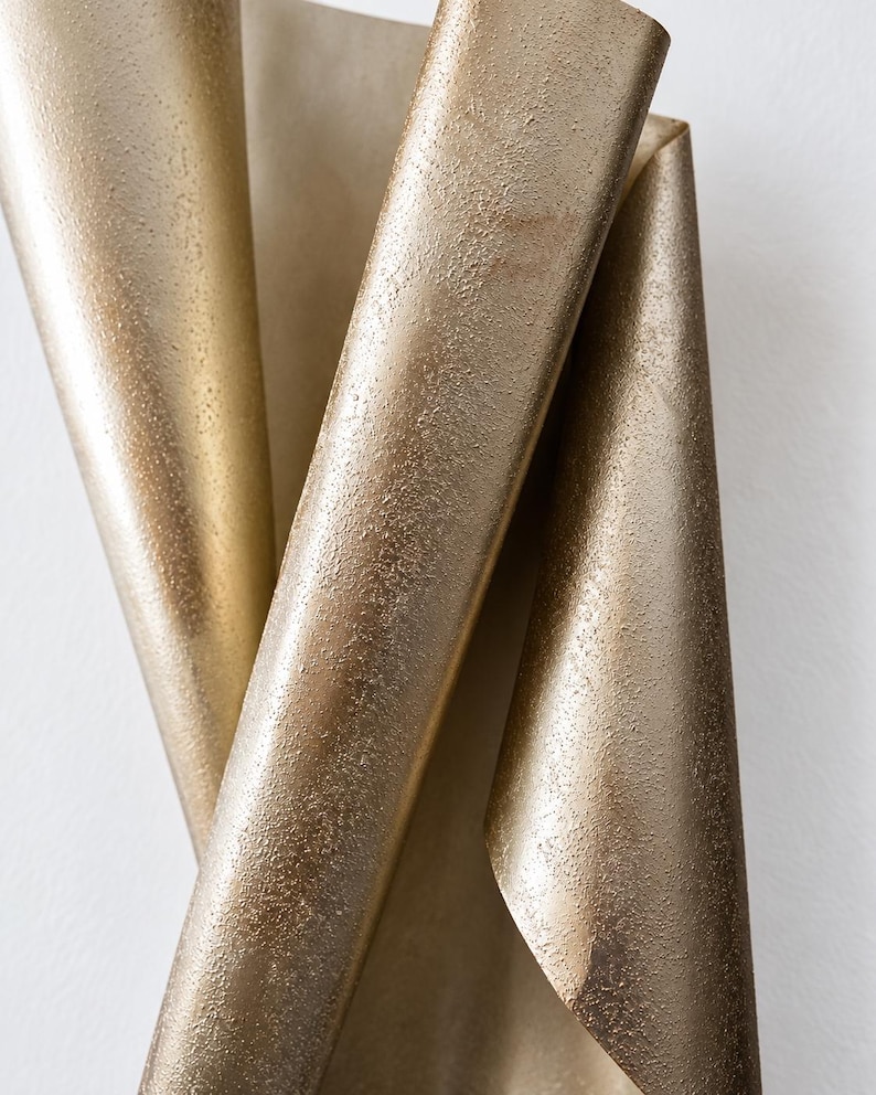May include: A sheet of metallic gold paper with a textured, granular surface, folded into a series of vertical, overlapping curves that create a dynamic, three-dimensional shape with varying light reflections across the uneven, shimmering finish.