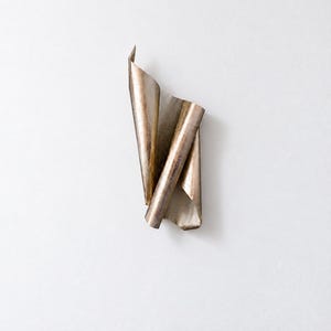 May include: A wall-mounted sculpture crafted from a single sheet of metallic material with a weathered, bronze-toned finish. The object features a series of vertical folds and curves, creating a layered, rolled appearance with sharp edges.