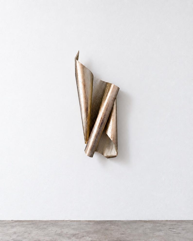 May include: A wall-mounted sculpture crafted from a single sheet of metallic material with a weathered, bronze-toned finish. The object features a series of vertical folds and curves, creating a layered, rolled appearance with sharp edges.