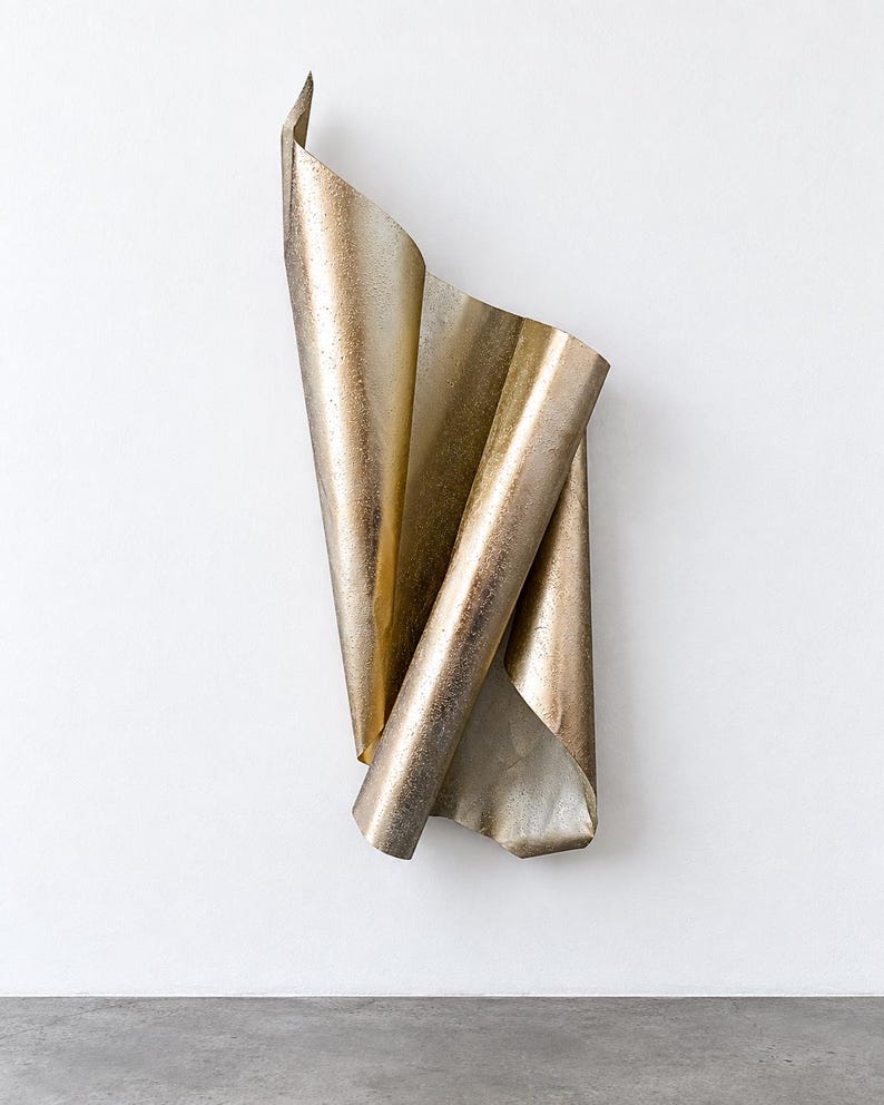 May include: A vertical wall sculpture made of a single sheet of metallic gold material, folded and curved into an abstract, flowing form. The surface features a textured, speckled finish with varying light reflections across its undulating contours.