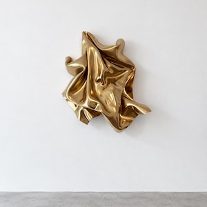 May include: A wall-mounted sculpture crafted from polished gold-toned metal, featuring a series of fluid, organic folds and deep curves that create a sense of movement and light reflection across its smooth, metallic surface.