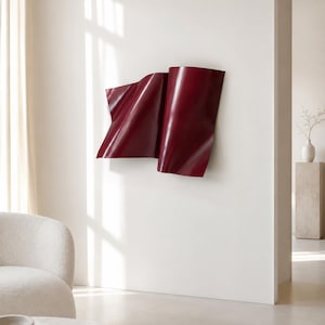 Könnte beinhalten: Eine tiefburgunderrote Wandskulptur mit einer glatten, geschwungenen und gewellten Oberfläche, die das Aussehen von gefaltetem Stoff nachahmt. Das Stück ist flach an einer schlichten weißen Wand montiert und erzeugt sanfte Schatten und Lichter.